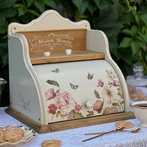 Può includere: Un portapane color crema con anta a serrandina decorato con illustrazioni di fiori e farfalle. L'unità include una base in legno e un ripiano superiore con la scritta La Maison Blanche stampata sul pannello posteriore e un pomello.