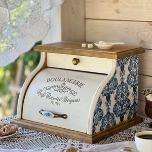 May include: A wooden bread bin with a roll-top door featuring the text BOULANGERIE Cafe Chaudet Beignets PARIS. The sides have a blue and white damask pattern, and the front includes a ceramic handle. The top is flat wood with a small white dish.