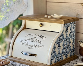 May include: A wooden bread box with a roll-top door featuring the text BOULANGERIE Cafe Chaudet Beignets PARIS. The sides have a blue and white damask pattern, and the front includes a ceramic handle. The top is flat wood with a small white dish.