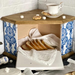May include: A corner bread bin with a natural wood top and base, featuring side panels decorated with a blue and white floral pattern. The interior contains sliced bread on a white cloth, while the top holds a white teacup and sugar cubes.