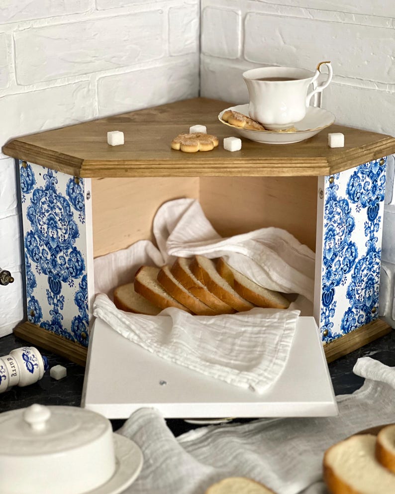 May include: A corner bread bin with a natural wood top and base, featuring side panels decorated with a blue and white floral pattern. The interior contains sliced bread on a white cloth, while the top holds a white teacup and sugar cubes.