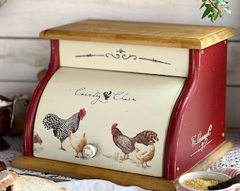 May include: A red and cream bread box with a roll-top door featuring illustrations of chickens and the text "Country Charm". The unit has a natural wood top and base, a small white ceramic knob, and decorative scrollwork accents on the front panel.