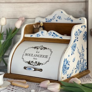 May include: A white wooden bread box with a roll-top door featuring the black text La BOULANGERIE Paris. The exterior is decorated with blue floral patterns on a white background, accented by a metal handle and a natural wood base.
