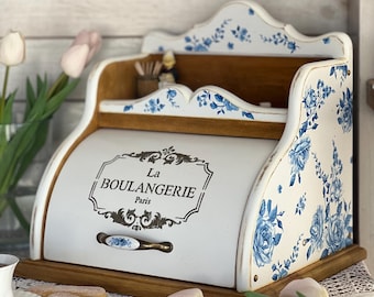May include: A white wooden bread bin with a roll-top door featuring the black text La BOULANGERIE Paris. The exterior is decorated with blue floral patterns on a white background, accented by a metal handle and a natural wood base.