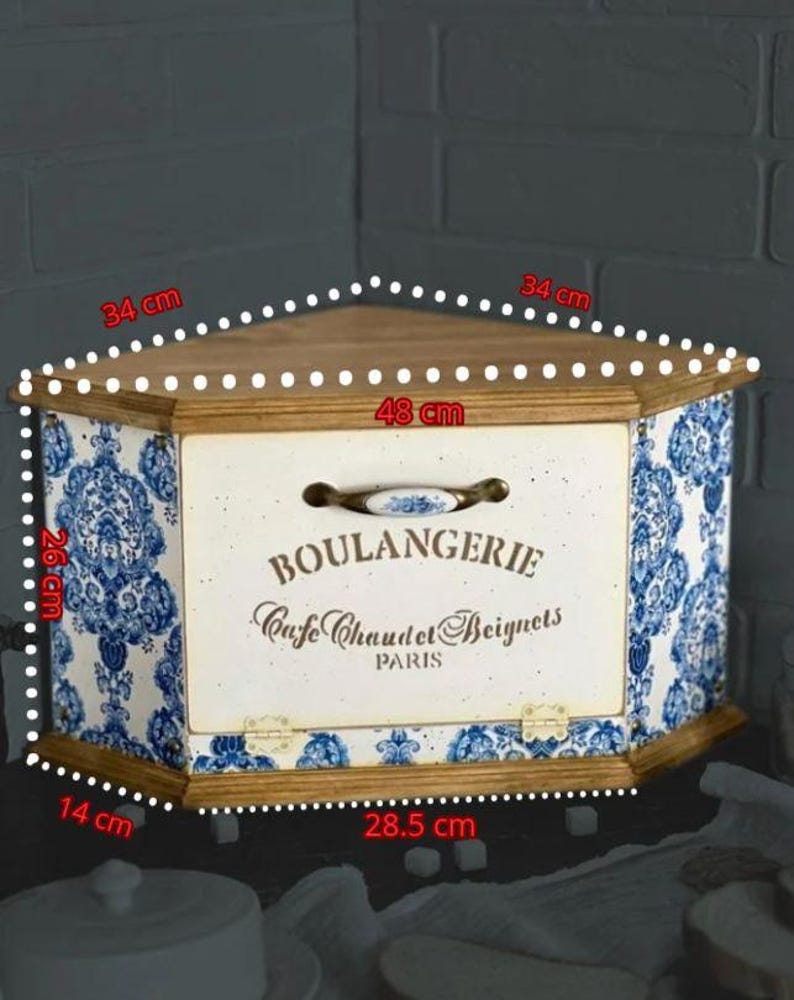 May include: A corner bread box with a wooden top and base, featuring blue and white floral patterns on the sides. The front panel is cream-coloured with the text BOULANGERIE, Cafe Chaud et Beignets, PARIS, and a handle. Dimensions are 34 cm, 48 cm, 26 cm, 14 cm, and 28.5 cm.