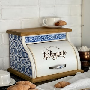 Puede incluir: Una panera blanca y azul con tapa y base de madera. La panera presenta patrones de baldosas florales azules en los laterales y un tirador decorativo. El texto "La Baguette" está impreso en la parte delantera. Una taza y un cruasán encima.