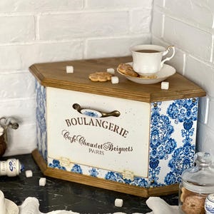 May include: A wooden bread bin with a white front panel featuring the text BOULANGERIE Cafe Chaud et Beignets PARIS and a blue and white floral pattern on the sides. The bin has a metal handle, hinges, and a flat top surface with a tea cup and biscuits.