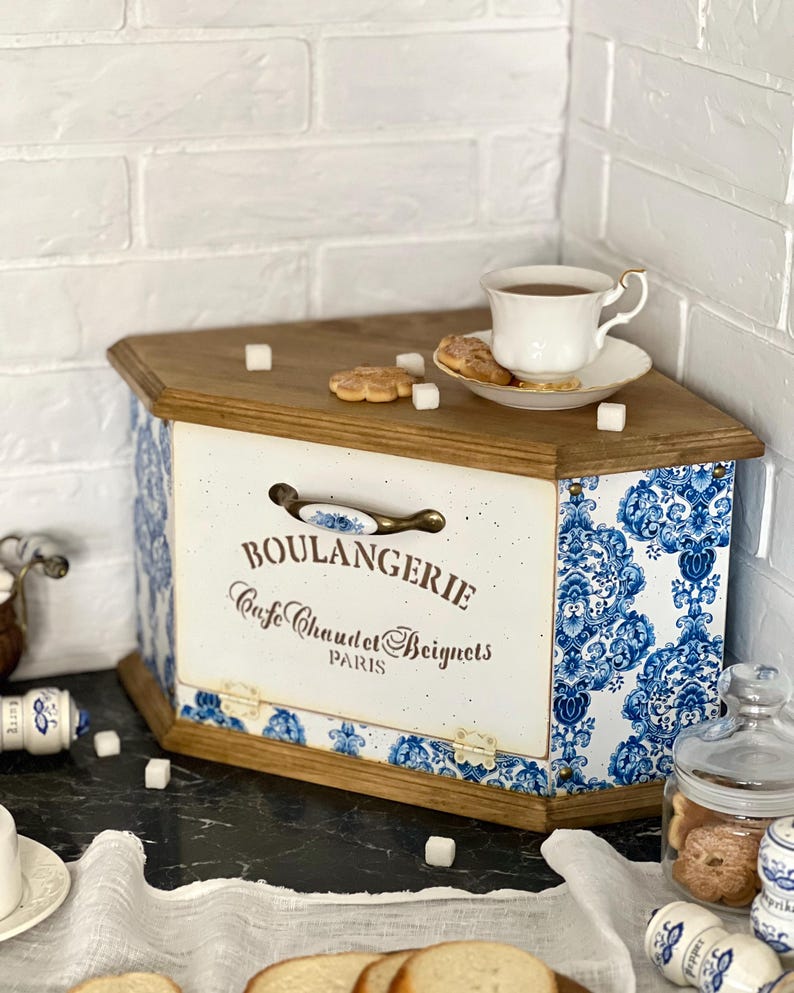 May include: A wooden bread bin with a white front panel featuring the text BOULANGERIE Cafe Chaud et Beignets PARIS and a blue and white floral pattern on the sides. The bin has a metal handle, hinges, and a flat top surface with a tea cup and biscuits.