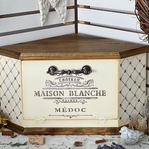 May include: A wooden corner storage chest with a dark brown top and base. The front and side panels are cream-coloured with a black lattice pattern. The front features a black metal handle and the text CHATEAU MAISON BLANCHE FRANCE MÉDOC.