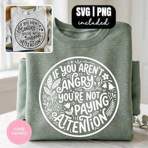 May include: Sage green sweatshirt with a white circular graphic that reads "If You Aren't Angry, You're Not Paying Attention." The design includes floral and leaf accents. A smaller image of a similar sweatshirt is in the upper left corner.