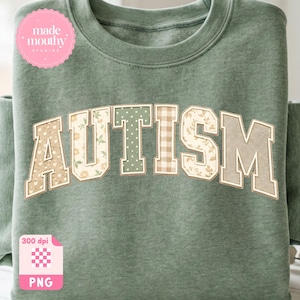Op de afbeelding: Saliegroene sweatshirt met het woord "AUTISM" in een collegestijl. De letters zijn versierd met bloemen-, stippen- en ruitpatronen. Een roze cirkelvormig logo met de tekst "made mouthy studios" staat linksboven.