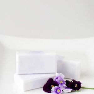 May include: Three bars of white soap with a hint of purple stacked on top of each other. A small bouquet of purple and white pansies are in front of the soap.