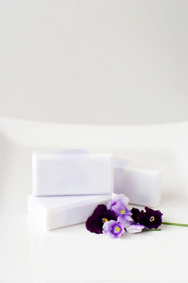 May include: Three bars of white soap with a hint of purple stacked on top of each other. A small bouquet of purple and white pansies are in front of the soap.