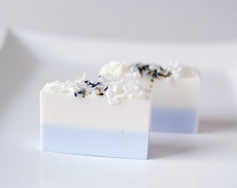 May include: Two bars of handmade soap with a white top layer and a light blue bottom layer. The soap is decorated with white coconut flakes and small blue and purple flower petals.