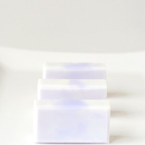 May include: Three bars of white soap with a subtle lavender hue. The soap bars are stacked on top of each other on a white surface.