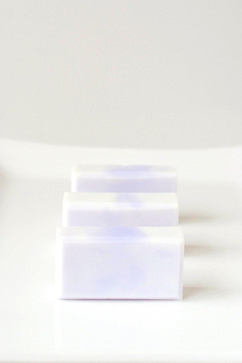 May include: Three bars of white soap with a subtle lavender hue. The soap bars are stacked on top of each other on a white surface.