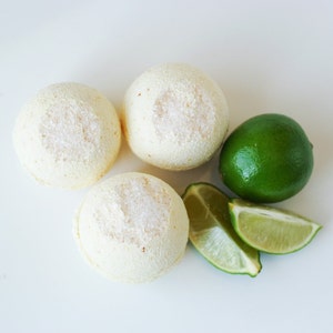 May include: Three round, off-white bath bombs with a textured surface are arranged with a whole green lime and two lime wedges. The items are set against a white background, creating a fresh, spa-like aesthetic.