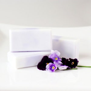 May include: Three bars of lavender-coloured soap with a delicate floral scent, arranged on a white surface with purple and white pansies.