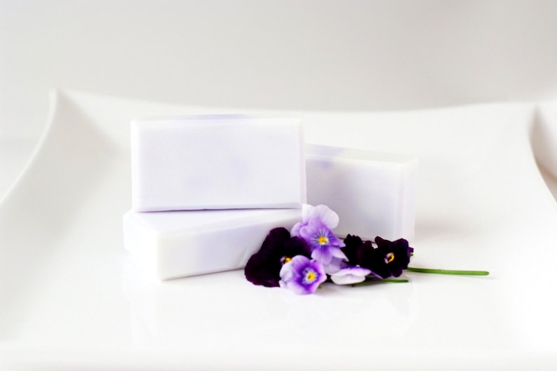 May include: Three bars of lavender-coloured soap with a delicate floral scent, arranged on a white surface with purple and white pansies.