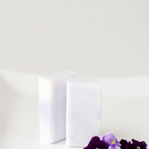 May include: Two white bars of soap with a purple hue, sitting on a white surface. A small bouquet of purple and white pansies are in the foreground.