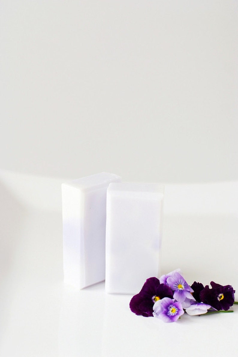 May include: Two white bars of soap with a purple hue, sitting on a white surface. A small bouquet of purple and white pansies are in the foreground.