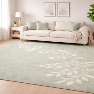 May include: A large, rectangular, light green rug with a white leaf design. The rug is in a living room setting with a white sofa, pillows, and a potted plant. The rug's dimensions are suitable for a medium to large living space.