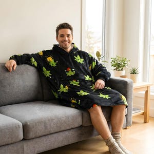 May include: A black hooded blanket featuring a pattern of green cannabis leaves and suns. The blanket has a large front pocket and is worn by a person on a grey sofa. The blanket is made of a soft, plush material.