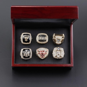 May include: A set of six championship rings displayed in a dark red wooden box. The rings feature gold and silver tones with intricate detailing, including the image of a bull. The box is open, revealing the rings set against a black interior.