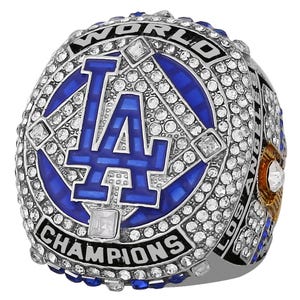 May include: A silver and blue championship ring featuring the letters "LA" in blue, encircled by white gemstones. The ring is inscribed with "WORLD CHAMPIONS" and "LOS ANGELES".