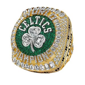 Puede incluir: Un anillo de campeonato dorado con la palabra "CELTICS" en letras blancas sobre un fondo verde, con un trébol adornado con diamantes. El anillo está incrustado con diamantes y tiene la palabra "CHAMPIONS" y años grabados en el lateral.