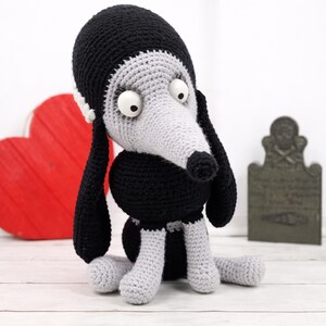 Peut inclure: Un jouet pour chien au crochet noir et gris. Le chien a de grands yeux blancs, de longues oreilles noires et un nez noir. Le jouet est sur une surface blanche avec un cœur rouge et une pierre tombale décorative en arrière-plan.