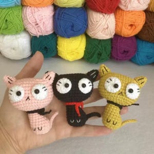 Peut inclure: Trois figurines de chat crochetées en rose, noir et jaune, chacune avec de grands yeux blancs et de petits détails noirs. Le chat noir porte une écharpe rouge. Le fond présente des pelotes de laine colorées, suggérant un thème artisanal.