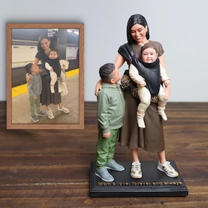 Op de afbeelding: 3D-geprinte sculptuur van een moeder en twee kinderen. De moeder houdt een baby vast in een draagzak. De sculptuur staat op een zwarte basis met de inscriptie "Mom's love: forever and always". Een ingelijste foto van dezelfde scène staat links.