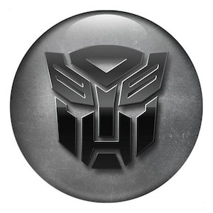 May include: A glossy, metallic grey and black Transformers logo emblem. The emblem is a stylized robot head, set against a textured grey circular background. The design features sharp angles and a reflective surface.