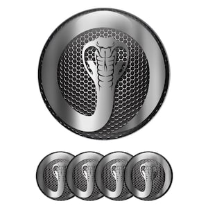 May include: A set of five silver-colored car emblems featuring a cobra design. The emblems have a circular shape with a mesh background and a glossy finish. The cobra is depicted in a stylized, detailed manner.
