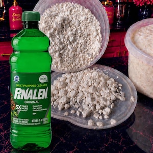 May include: A green plastic bottle of Pinalen Original multipurpose cleaner with a dark green cap, featuring a label with white text and a pine cone graphic. Next to it are two clear plastic containers filled with white, crumbly, solid material.