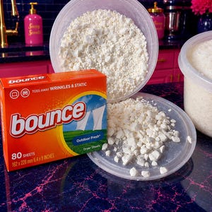 Puede incluir: Una caja naranja de hojas para secadora Bounce que indica 80 hojas, 162 x 228 mm, 6,4 x 9 pulgadas, junto a un recipiente de plástico transparente lleno de trozos sólidos, blancos y quebradizos. Algunos trozos están esparcidos en la tapa.