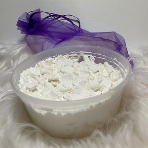 May include: A clear plastic container filled with white, irregularly shaped pieces. A sheer purple drawstring bag is behind the container. The container sits on a fluffy white surface.