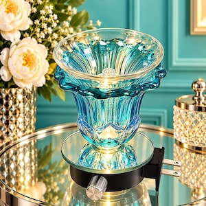 May include: A plug-in wax warmer featuring a translucent, scalloped-edge blue glass bowl. The base is black with a silver control knob and a standard electrical plug. The glass bowl is illuminated from within, casting a warm glow.
