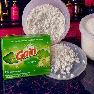 May include: A green Gain Original dryer sheets box with 80 sheets measuring 6.4 x 9 inches, positioned next to a clear plastic container filled with white, crumbled, solid material. The box features the Gain logo and text, Seriously Good Scent.