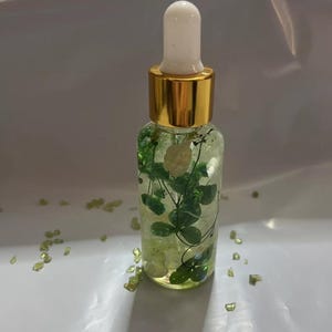 May include: A clear glass bottle with a gold cap and white dropper filled with green botanicals and crystals. The bottle contains a liquid and is surrounded by small green crystals.