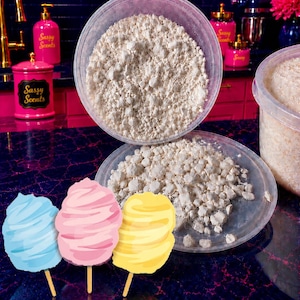 May include: A clear plastic container filled with small, off-white, irregular wax granules, with a matching lid resting below it. Three graphic illustrations of cotton candy on sticks in blue, pink, and yellow are positioned in the foreground.