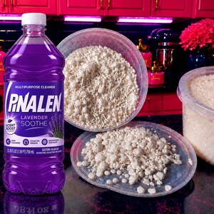 May include: A clear plastic bottle of Pinalen Lavender Soothe multipurpose cleaner with purple liquid, featuring a white label with text, next to a clear plastic container and lid filled with a textured, off-white granular substance.
