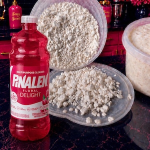 May include: A bottle of red Pinalen Floral Delight multipurpose cleaner with a white cap and label, positioned next to a clear plastic container filled with white, crumbly, solid material and a matching lid covered in the same white substance.