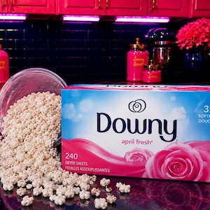 May include: A rectangular box of Downy April Fresh dryer sheets featuring a pink rose graphic and the text 240 Dryer Sheets. Next to the box is a clear plastic container tipped over, spilling small, white, flower-shaped laundry scent beads.