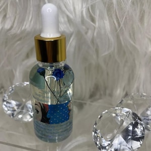 May include: A clear glass bottle with a white dropper and gold cap, filled with a light blue liquid and blue flowers. The bottle has a label with text and illustrations. Decorative crystal accents are in the foreground.