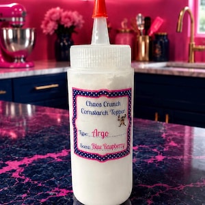 May include: A clear plastic squeeze bottle with a red pointed cap, filled with white powder. The label reads Chaos Crunch Cornstarch Topper, Type: Argo, and Scent: Blue Raspberry, featuring a small cartoon character graphic on a blue and white polka dot border.