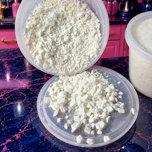 May include: A clear plastic container filled with small, irregular, off-white solid chunks. The container is tilted, spilling some of the white granular material onto its circular, translucent plastic lid, which rests flat below the main container.