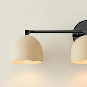 Puede incluir: Aplique de pared moderno de dos luces, con estructura de metal negro y pantallas en forma de c&uacute;pula de color crema. La l&aacute;mpara est&aacute; montada sobre una base circular negra. Las luces est&aacute;n encendidas.