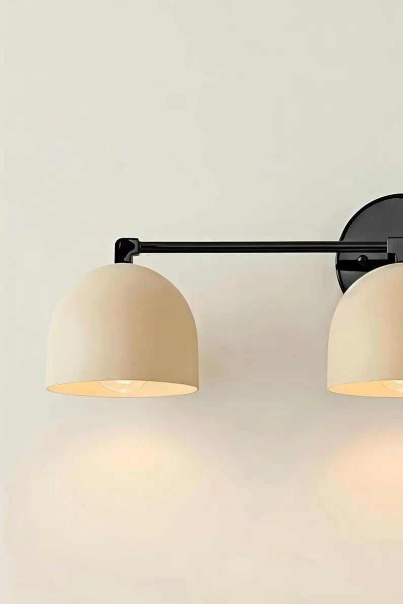 Puede incluir: Aplique de pared moderno de dos luces, con estructura de metal negro y pantallas en forma de c&uacute;pula de color crema. La l&aacute;mpara est&aacute; montada sobre una base circular negra. Las luces est&aacute;n encendidas.